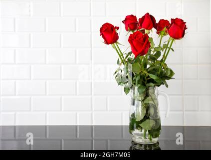 Beautiful red roses in a glass jug on black granite kitchen counter and white tile background. Stock Photo