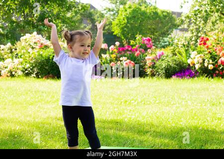 Little cute smiling girl 1-3 in a white T-shirt stands with raised hands on the grass against the background of flowers Stock Photo