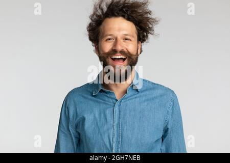portrait of happy curly hair guy in denim shirt with long beard and moustache having fun and laughing on grey background in studio Stock Photo