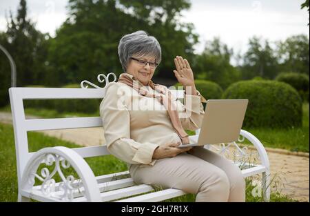 Happy senior woman sitting on park bench, video calling family and waving hello at laptop screen Stock Photo