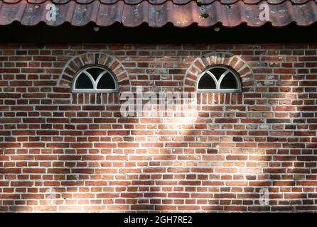 An old bricked wall with two arched windows and a tiled roof. Seen in the Netherlands Stock Photo