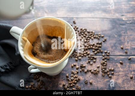 Pouring boiling water on ground coffee in a paper filter and a porcelain holder on a mug for an aromatic brewed hot drink, rustic wooden table with so Stock Photo