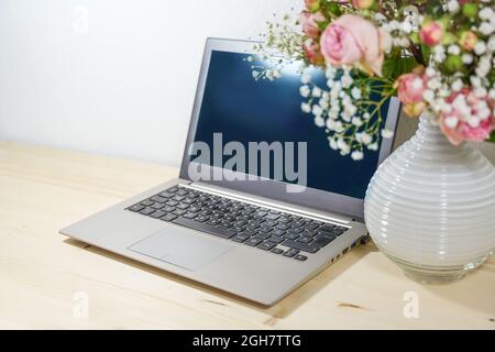 Silver gray laptop with a dark screen on a light wooden table and a vase with flower bouquet in the foreground, copy space, selected focus, narrow dep Stock Photo