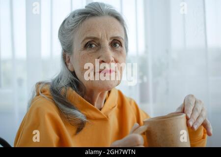 Dreamy eccentric middle aged woman sitting with cup of black tea or coffee, looking at camera. Peaceful mature lady enjoying no stress calm positive Stock Photo