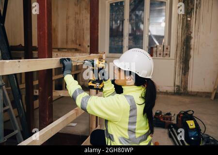 Female worker in hardhat using drill on wooden plank while working at construction site Stock Photo