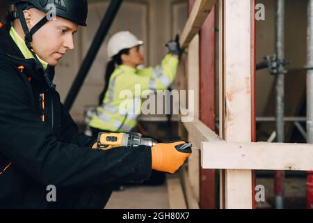 Side view of male worker using drill on wooden plank while working at construction site Stock Photo