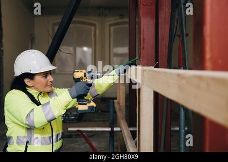 Female worker using drill on wooden plank while working at construction site Stock Photo