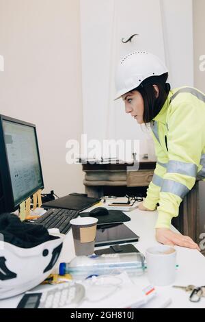 Female building contractor in hardhat using computer while leaning on desk at office Stock Photo