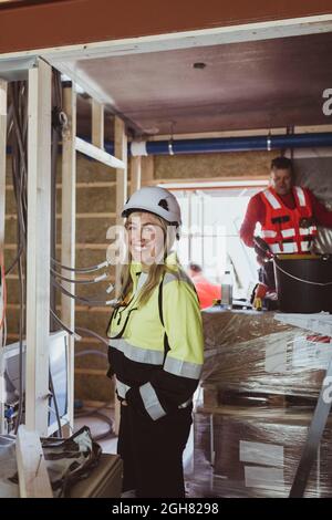 Happy female building contractor standing with hand in pocket while male construction worker working at site Stock Photo