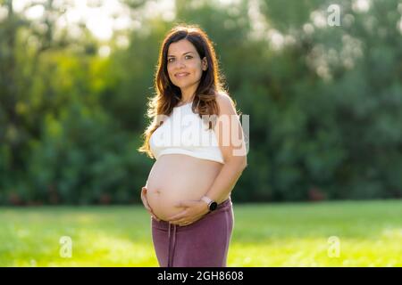 Pregnant woman touching her belly while looking at the camera outdoors Stock Photo