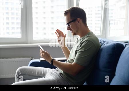 Happy handsome guy is waving hello into phone webcam, smiles and greeting. Video call, video conference, online meeting. Side view at young man during virtual meeting on the smartphone Stock Photo