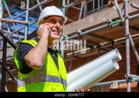 Male builder foreman, worker or architect on construction site, happy, smiling, holding building plans and talking on his cell phone Stock Photo