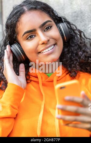 Outdoor portrait of beautiful happy mixed race African American girl teenager female young woman wearing an orange hoodie listening to music on wirele Stock Photo