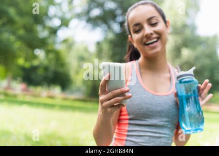 happy and blurred woman holding sports bottle with water and using smartphone in park Stock Photo