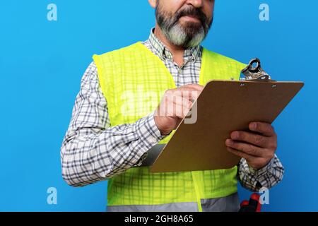 Male builder or handyman in uniform holding clipboard against blue background Stock Photo
