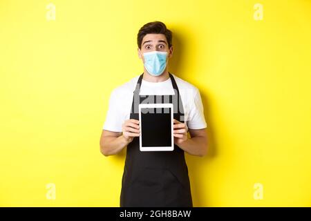 Concept of covid-19, small business and pandemic. Surprised waiter in black apron and medical mask showing tablet screen, standing over yellow Stock Photo
