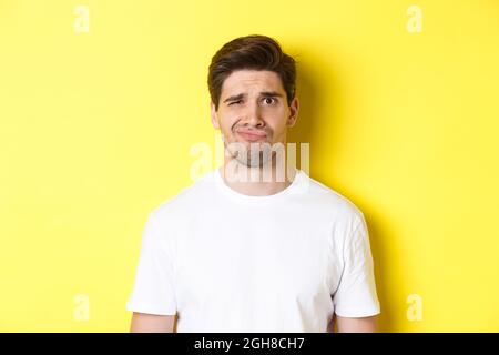 Close-up of dispelased young man in white t-shirt looking doubtful, grimacing unsatisfied, standing over yellow background Stock Photo