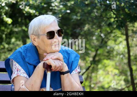 Happy old woman in sunglasses sitting with walking cane on a bench in park. Life in retirement, enjoying the nature Stock Photo