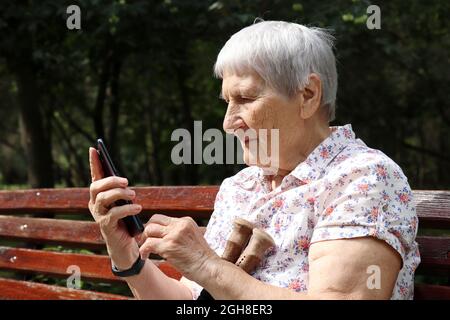 Elderly woman sitting with smartphone on a bench in park. Online communication, sms, life in retirement Stock Photo
