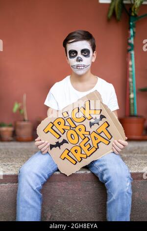 Teenage boy with face characterized as a skull sitting outdoors. He is serious and holds in his hands a sign with the text Trick or Treat. Halloween. Stock Photo