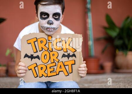 Close up of boy with face painted like a dead man holding a sign with the text Trick or Treat. He is outdoors. Halloween concept. Stock Photo