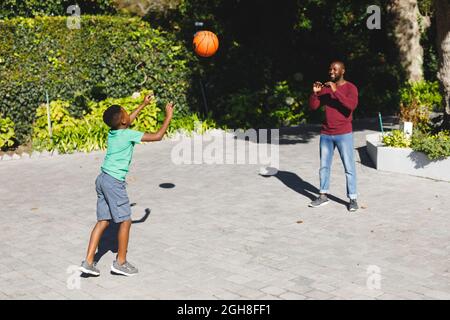 African american father with son smiling and playing basketball in garden