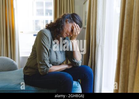 Stressed senior caucasian woman sitting on sofa by window holding head Stock Photo