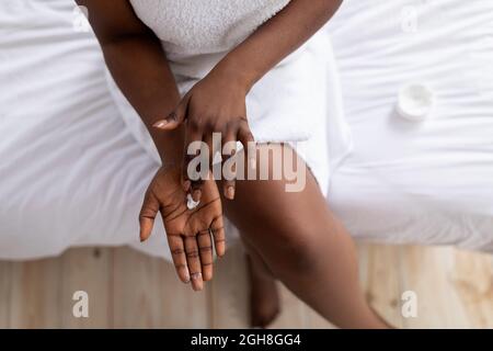 Top view of young black lady applying hand cream, sitting on bed after bath, wearing towel, pampering herself at home Stock Photo
