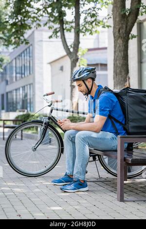 Side view of muslim courier in blue t-shirt and thermo backpack using cellphone on bench near bike Stock Photo