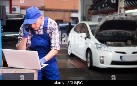 Auto mechanic with laptop talking on mobile phone in auto repair shop Stock Photo