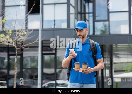 Arabian courier in uniform using cellphone and holding takeaway drink on urban street Stock Photo