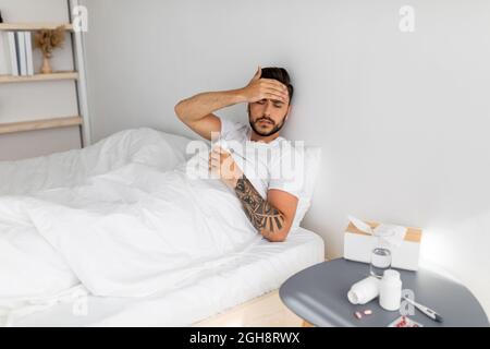 Feeling unwell. Ill man having severe headache suffering from pain and touching forehead, sitting in bed, copy space Stock Photo
