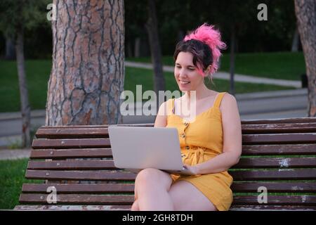 Young caucasian woman with pink hair working on a laptop in a park bench. Stock Photo