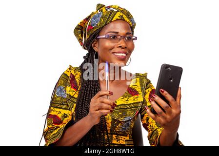 happy beautiful african mature woman sitting making a video call in her mobile telepone holding her pen in hand Stock Photo
