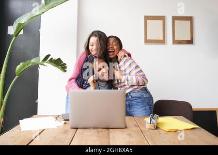 A group of women hugging each other euphorically celebrate the good news in front of the computer. Successful multiethnic group excited and happy. Stock Photo