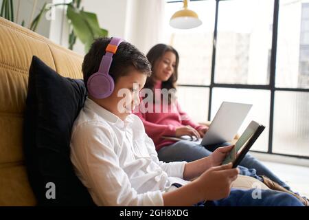 Boy with headphones playing with a tablet on the sofa at home while his mother works remotely on her laptop. Concept of remote work and family Stock Photo