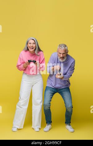 KYIV, UKRAINE - AUGUST 10, 2021: full length of happy interracial and mature couple playing video game on yellow Stock Photo