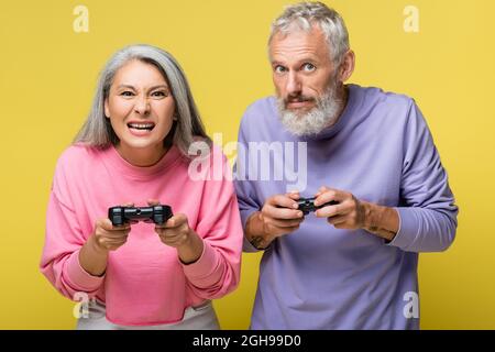 KYIV, UKRAINE - AUGUST 10, 2021: tensed interracial and mature couple playing video game isolated on yellow Stock Photo