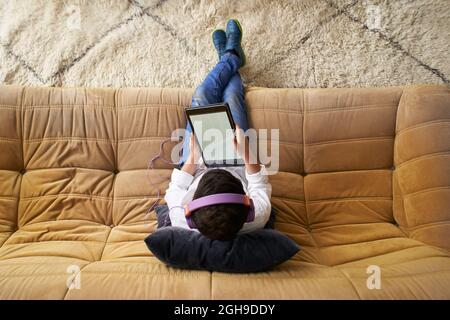 Top view of a caucasian boy with helmets playing with a tablet sitting on the sofa at home. Stock Photo