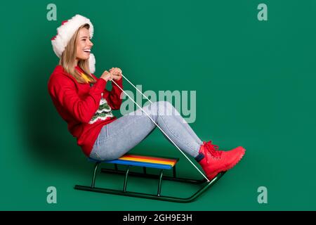 Full length profile photo of crazy santa young lady slide on sled wear sweater jeans boots isolated on green background Stock Photo