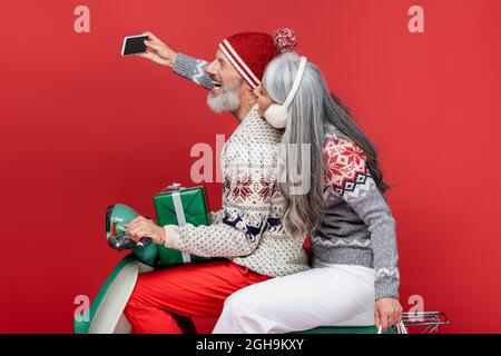happy and mature interracial couple in sweaters taking selfie while riding motor scooter on red Stock Photo