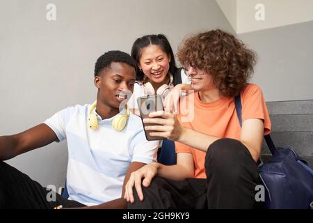 Group of teenage students of different ethnicities sitting outside on the stairs of the university using the cell phone. Stock Photo