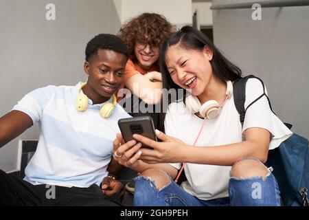 A group of teenage students of different ethnicities laugh while watching something on a mobile phone sitting outside on the stairs of the university. Stock Photo