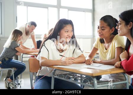 Smiling students working together as a team in class. In the background the teacher helps other classmates. Stock Photo