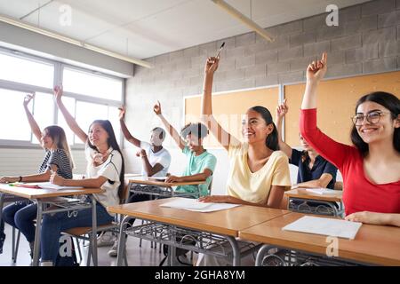 Multi-ethnic students in the classroom with hands raised. Smiling young people answering teacher's question. Stock Photo
