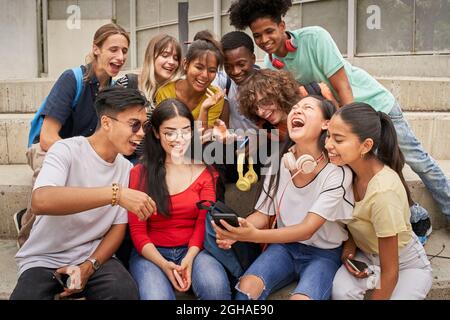 Multi-ethnic group of students using mobile phone and laughing. Teenagers using a smart phone, surfing the web. Having fun together. Stock Photo