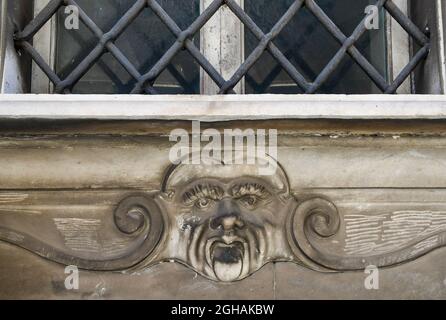 Detail of an old building with a bas-relief representing the frowned face of a man under a window closed by an iron grate, Livorno, Tuscany, Italy Stock Photo