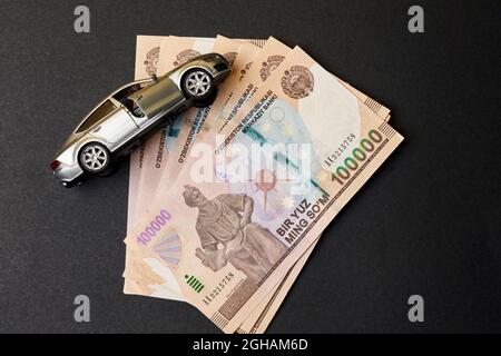 Uzbek sums and car toy. Concept of auto vehicle insuarance in Uzbekistan. Pile of 100000 sums and modern automobile toy on black background. Auto tax Stock Photo