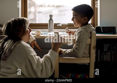Smiling son talking with mother while sitting on chair at home Stock Photo