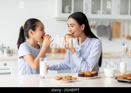 Dessert time for sweet tooth. Happy asian girl and young woman eating cookies, sitting at dinner table in kitchen Stock Photo